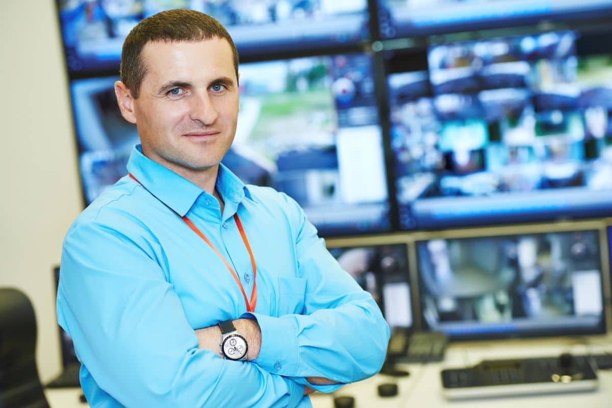man in blue shirt with arms crossed standing in front of a group of security monitors